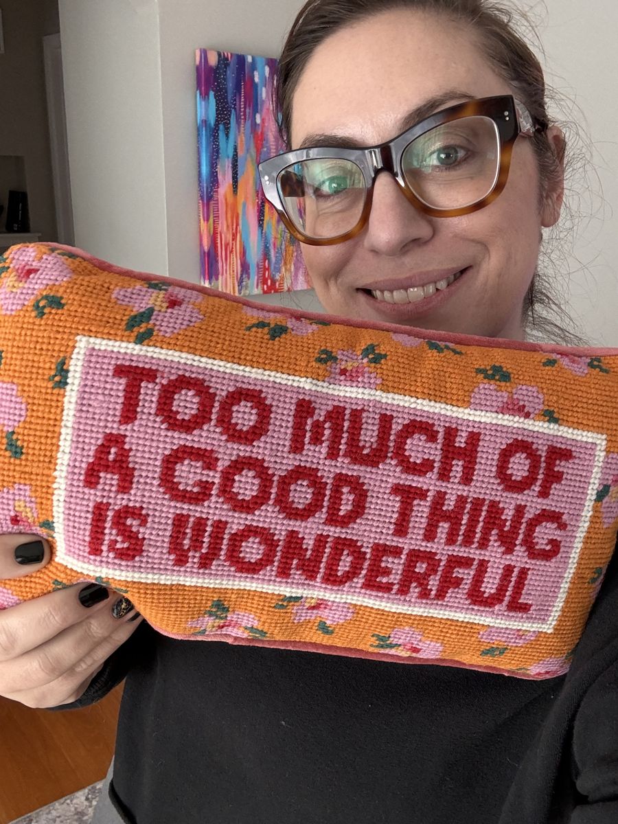 Laura smiling and holding a needlepoint pillow that reads Too Much of a Good Thing Is Wonderful